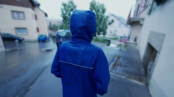Back of child wearing raincoat walking in street in rainy day. Kid wears blue waterproof coat