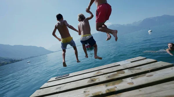 Family jumping into water father and young boys running together on pier diving into lake