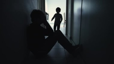 Depressed father being consoled by son sitting in corridor in silhouette. Child hugging sad parent during crisis