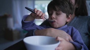 Cute little boy eating cereal on mother lap. Authentic real life domestic lifestyle family scene of child wearing pajamas bonding with mom at home