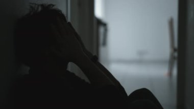 Sad unhappy man sitting on floor holding his forehead while having mental pain. Depressed person in dark corridor with low light environment. Dramatic scene