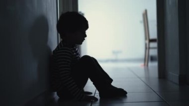 Sad child suffering from depression sitting alone in corridor feeling loneliness. Scared fearful small boy covering face in silhouette at home