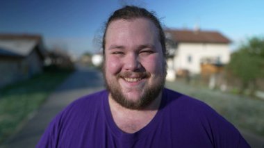 One joyful young chubby person stands outdoors smiling at camera. Overweight male person closeup face
