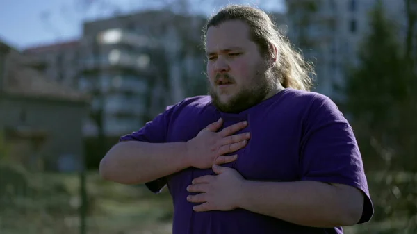 One young overweight man having chest pains standing outdoors. A fat person feeling stressful pain with difficulty in breathing