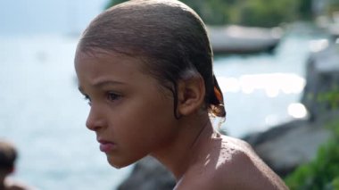 One wet contemplative young boy drying in the sun outside after bathing at lake. Profile of serious pensive child