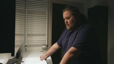 Sad young man standing in bathroom concerned about problems with pensive expression. A male overweight person in front of mirror