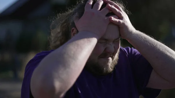 One frustrated overweight young man standing outdoors feeling regret. Anxious upset person hitting himself in the head in anguish due to failure