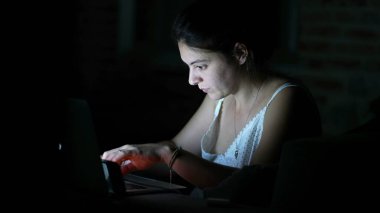 Gecenin karanlığında dizüstü bilgisayarın önünde interneti karıştıran konsantre bir kadın. Pijamalı bir kadın akşamları bilgisayar ekranına odaklanmış bir ifadeyle bakıyor.