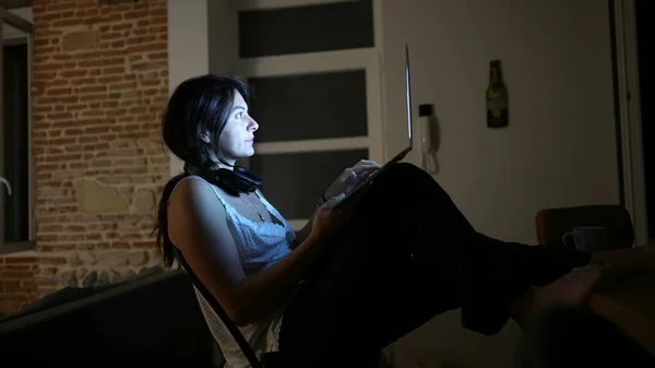 Woman working at night at home in front of laptop computer in the dark. Computer screen glowing on person face while browsing internet online