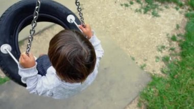 Child seated on tire swing spinning at playground park daydreaming. Top view perspective of one small boy spins