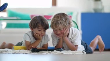 Candid of two young boys watching media on tablet screen lying on floor at living room floor at home. Children staring at technological device indoors