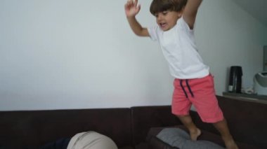 Active child jumping on mother at couch. Funny family authentic scene. Mom grimacing in slow motion lying on sofa. Parent pain emotion