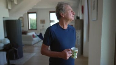One thoughtful senior man standing at home living room looking out window with pensive expression. 70s male older mature person holding cup of coffee in morning