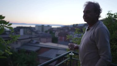 Mature man standing outside looking at scenic view while holding glass in sunset time. Retired senior older male person in 70s enjoying retirement