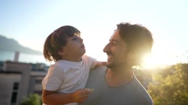 Child son kissing father many times. Parent holding kid standing outdoors in sunset time. Love and fatherhood concept