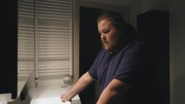 Sad young man standing in bathroom concerned about problems with pensive expression. A male overweight person in front of mirror