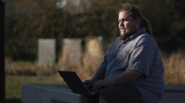 Pensive young man in front of laptop sitting outdoors in city park. Thoughtful freelancer working remotely using computer in tracking shot with movement