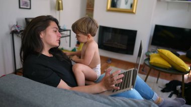 Mother trying to work and multi task with baby toddler at home sofa living room. Child bothering parent while working with laptop computer