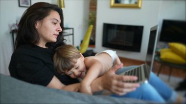 Mother trying to work and multi task with baby toddler at home sofa living room. Child bothering parent while working with laptop computer