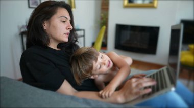 Mother trying to work and multi task with baby toddler at home sofa living room. Child bothering parent while working with laptop computer