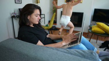Woman working from home using laptop with child jumping from sofa. Parent in front of computer screen browsing internet online multi tasking