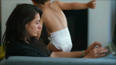 Mother working from home with small boy playing in background. Businesswoman in front of laptop computer works laying on couch. Modern motherhood lifestyle