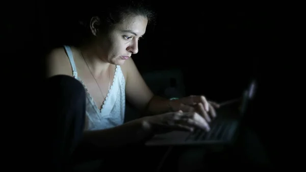 Woman working at night at home in front of laptop computer in the dark. Computer screen glowing on person face while browsing internet online