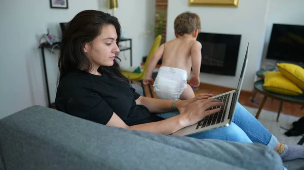 Woman working from home using laptop with child jumping from sofa. Parent in front of computer screen browsing internet online multi tasking