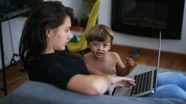 Mother parenting with child in front of laptop screen working from home. Parent sitting on couch with toddler boy looking at computer