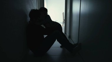 Depressed father being consoled by son sitting in corridor in silhouette. Child hugging sad parent during crisis
