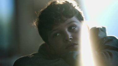 One contemplative kid daydreaming close up face. Pensive preteen kid leaning on table with arms crossed thinking with lens flare artistic reflection