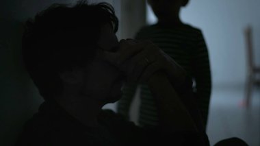 Sad father being held by son. Dramatic scene of male parent suffering sitting in corridor in the dark. Child consoling dad