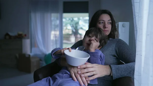 Cute little boy eating cereal on mother lap. Authentic real life domestic lifestyle family scene of child wearing pajamas bonding with mom at home