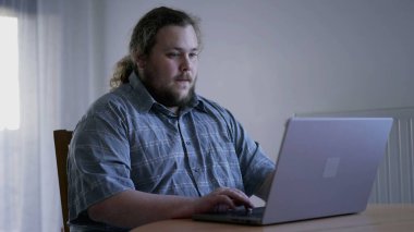 One casual overweight young man in front of laptop computer browsing internet at home