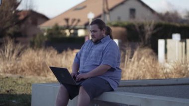 One thoughtful chubby student in front of laptop sitting outdoors in city bench. Pensive young man having an idea in front of computer. Eureka moment