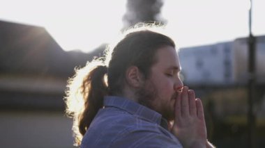 Spiritual young man praying to GOD outdoors in sunlight. A male overweight caucasian person with hands clenched in prayer. Hope and Faith concept