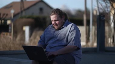 Focused young man using laptop sitting outside in city bench. Person working remotely in open space in front of computer screen outdoors