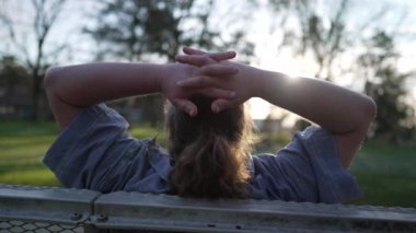 Back of a relaxed young man sitting at park bench enjoying nature and putting arms behind head resting outdoors