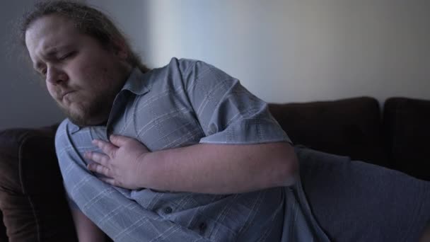 胸の痛みに苦しむソファの上に寝そべって健康上の問題を抱えている人 心臓病を患っている若い脂肪の男 心配な感情 — ストック動画