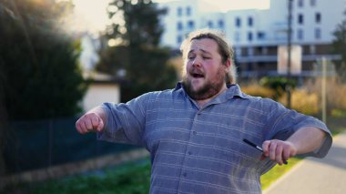 Happy man celebrating success holding phone and walking outdoors dancing in excitement outside in sunlight. One overweight guy feeling excitement