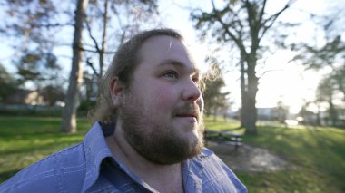 One happy chubby young man standing outdoors at park enjoying nature. Male caucasian person in contemplation
