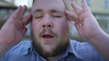 Portrait of person suffering from migraine pressing fingers to temples closing eyes with helpless face expression. One chubby young man having headache and stress