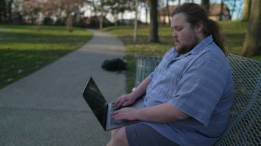 Person using computer outside seated at park bench. Millennial young man in 20s typing on laptop keyboard studies remotely