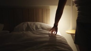 Young man laying in bed at night going to sleep. Male person lays under bedsheets and turning bedside night light off