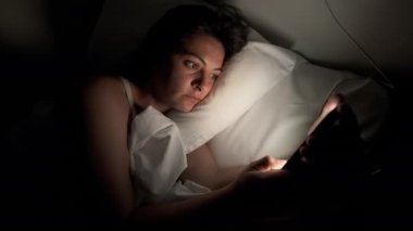 Woman laid in bed looking at smartphone screen with glowing light on face. Person using phone at night before sleep