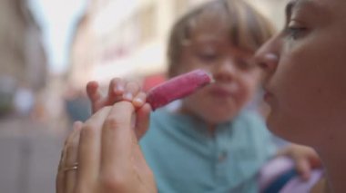 Mother sharing ice cream with son during summer day. Woman eating sweet dessert snack holding child in street while snacking