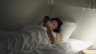 Woman looking at cellphone device laying in bed at night. Female person staring at glowing screen in the dark before bedtime