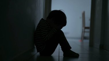 Small child covering face feeling despair. One small boy sitting in dark hallway corridor at home suffering alone in crisis