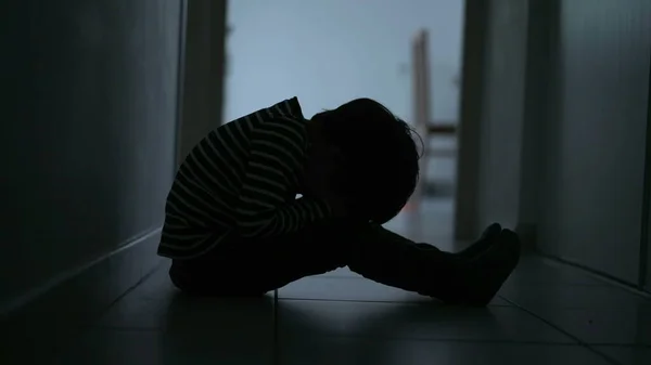 One scared desperate child covering face in fear sitting in dark corridor at home. Childhood loneliness and despair concept