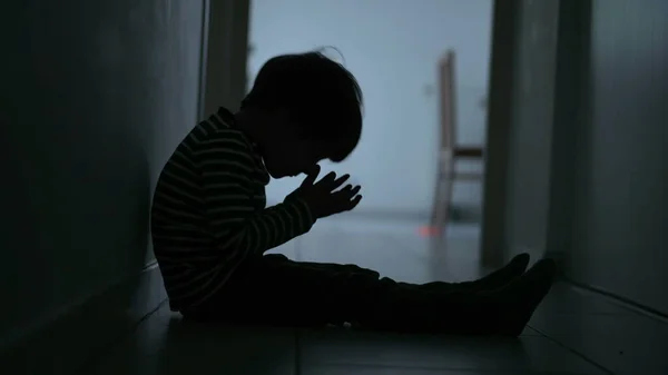 One scared desperate child covering face in fear sitting in dark corridor at home. Childhood loneliness and despair concept